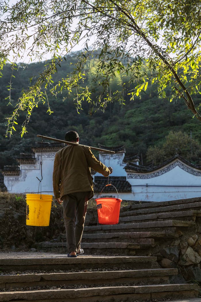 Back view faceless male in worn out clothes carrying plastic buckets with clean water by using shoulder pole while walking up on shabby street stairs in countryside