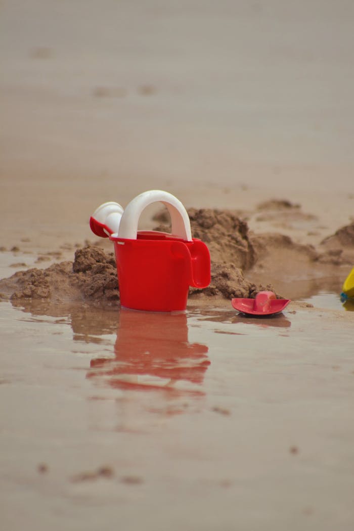 Red bucket and spade set on wet sand, hinting at a playful beach day.