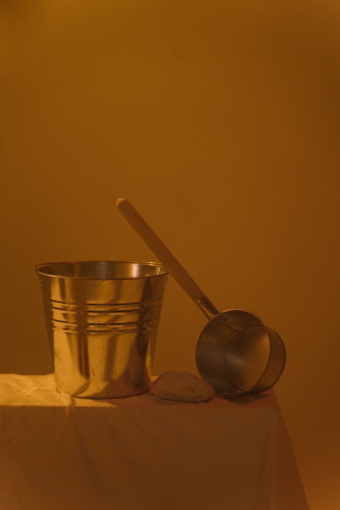A stainless steel bucket, ladle, and white cloth on table with warm lighting.
